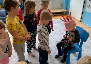 4 urodziny Wojtusia. Chłopiec siedzi na krzesełku. Dzieci zgromadzone wokół jubilata składają życzenia. Wojtuś ma na głowie czapkę w kształcie tortu.